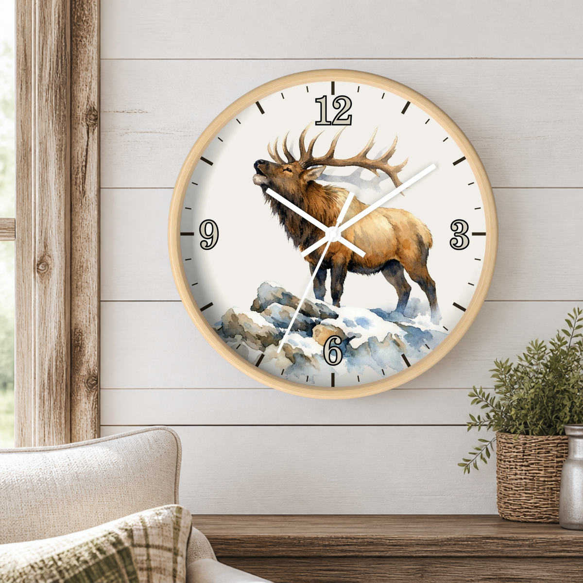 A round wall clock with a light wood frame hangs on a white shiplap wall above a wooden mantel. The clock face features a detailed illustration of a brown elk standing on snowy rocks, with black Arabic numerals and white hands.