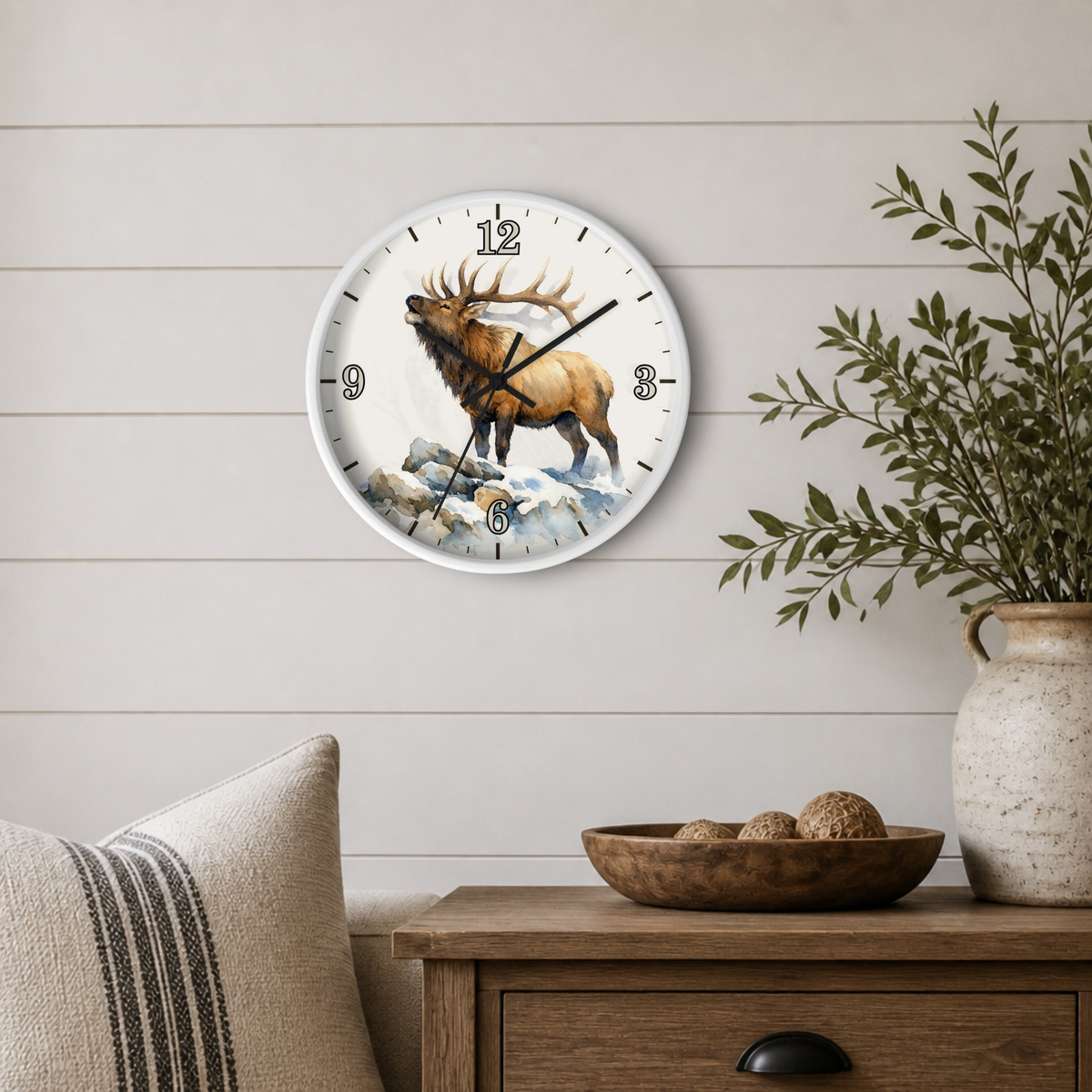 A round wall clock with a white frame and a detailed illustration of a brown elk standing on snowy rocks, mounted on a light beige shiplap wall above a wooden side table with a plant and decorative bowl.