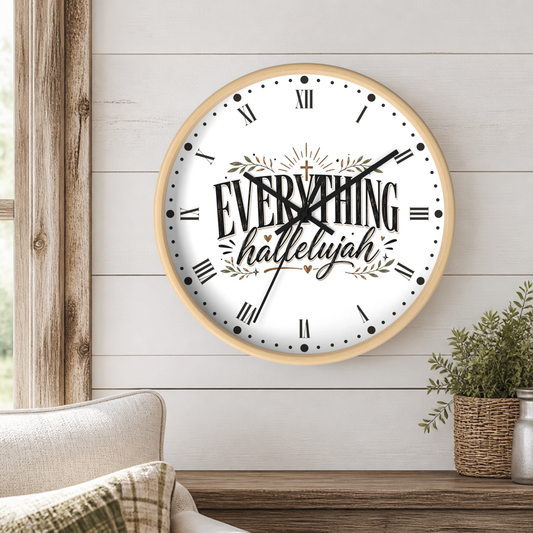 A round wall clock with a light wood frame hangs on a white shiplap wall above a wooden console table. The clock face is white with black Roman numerals, black hands, and the words 'EVERYTHING hallelujah' in decorative script and serif fonts, accented with small olive-green leaf motifs.
