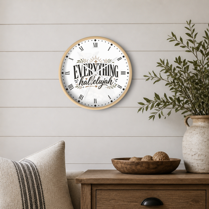 A round wall clock with a light wood frame and the words 'EVERYTHING halleyyah' printed on the white face, styled on a light wood shiplap wall above a wooden side table with a plant and pillows.