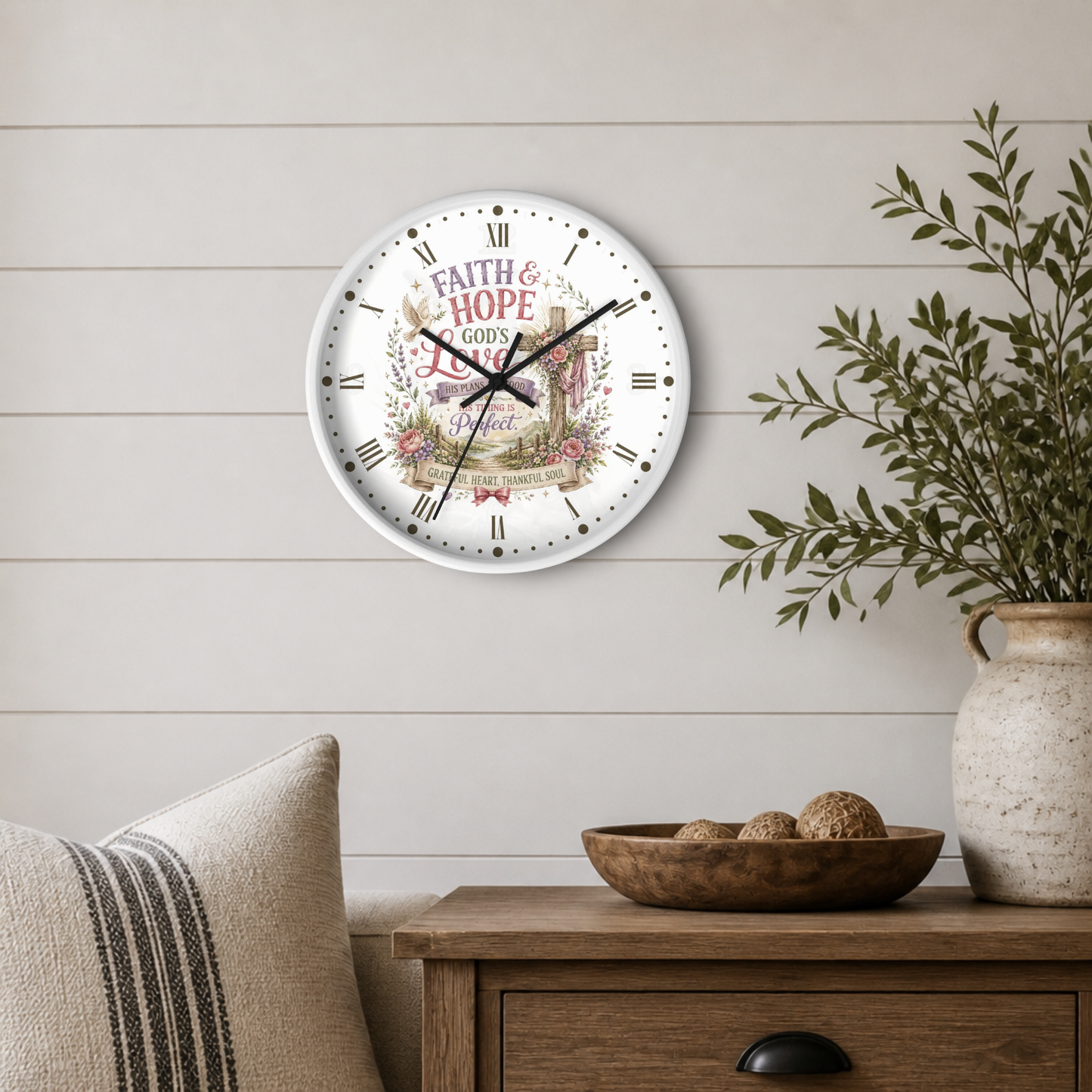 A round wall clock with a white face and black Roman numerals, featuring the words 'FAITH & HOPE & LOVE' and a floral illustration, styled on a light wood-paneled wall above a wooden side table with a plant and decorative bowl.