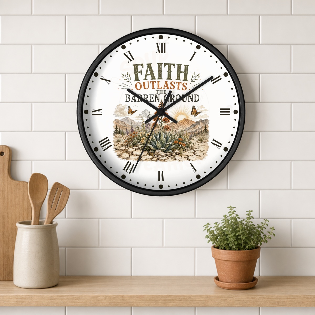 A round wall clock with a black frame and a white face featuring the text 'FAITH OUTLASTS THE BARREN LAND' above a desert landscape illustration, mounted on a white subway tile wall above a wooden shelf with kitchen utensils and a potted plant.