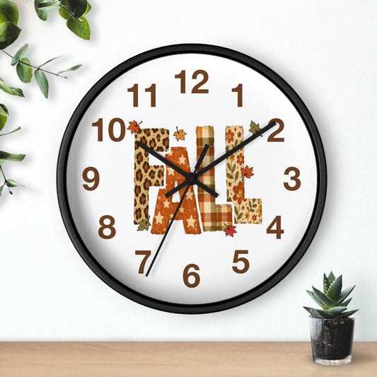 A round wall clock with a black frame and a white face, featuring the word "FALL" in autumn-themed lettering. The letters are filled with leopard print, orange plaid, and maple leaf patterns, with small brown numbers and black hands.