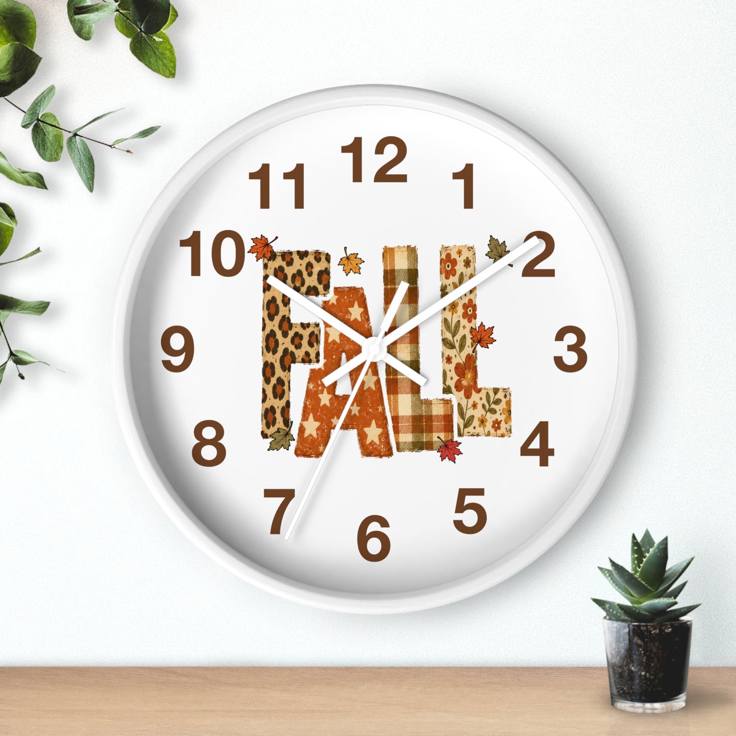 A round white wall clock with a fall-themed 'FALL' letter design in leopard print and plaid patterns, styled on a light wood surface against a white wall with small green plants.