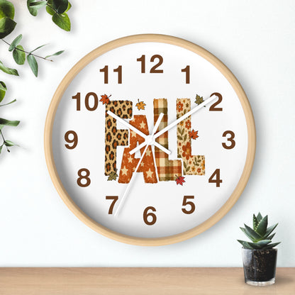 A round wall clock with a light wood frame and a white face, featuring the word "FALL" in autumn-themed letter patterns: leopard print, plaid, and solid brown. The clock is shown hanging on a white wall above a wooden surface, with small green plants in the corners.