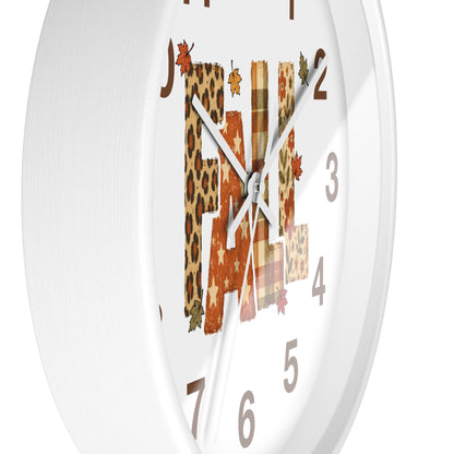 A white round wall clock with a decorative autumn-themed face. The letters of the word 'FALL' are filled with leopard print, orange stars, and plaid patterns, accented with small leaf graphics.