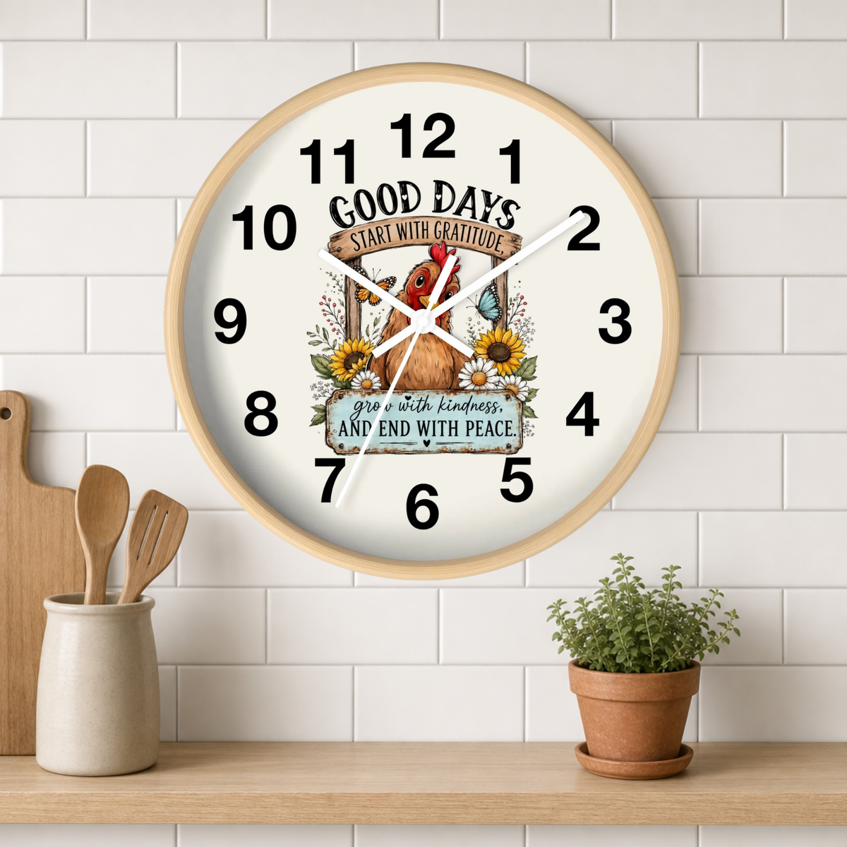 A round wall clock with a light wood frame hangs on a white subway tile wall above a wooden shelf. The clock face is white with black numerals and hands, featuring a central illustration of a brown rooster, sunflowers, and the text 'GOOD DAYS START WITH GENTLEMAN' and 'grace with kindness, and END WITH PEACE'.
