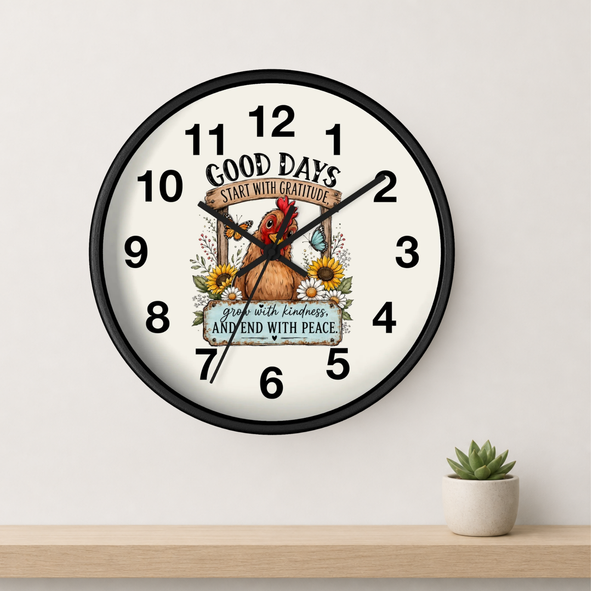 A round wall clock with a black frame and white face, featuring a rustic chicken illustration and motivational text. The clock is mounted on a light gray wall above a wooden shelf with a small potted succulent.