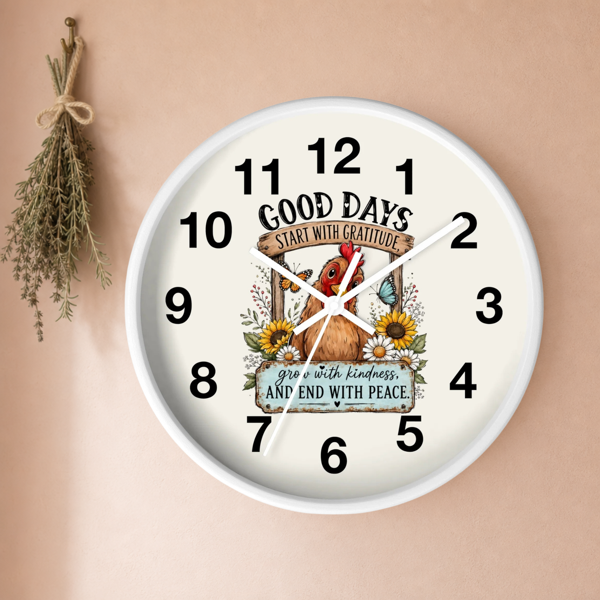 A round wall clock with a white frame and cream face, featuring a decorative autumn-themed illustration of a rooster, sunflowers, and leaves, with the text 'GOOD DAYS START WITH GRATITUDE' and 'grow with kindness, AND END WITH PEACE.' The clock is mounted on a warm beige wall with a small dried herb sprig hanging to the left.
