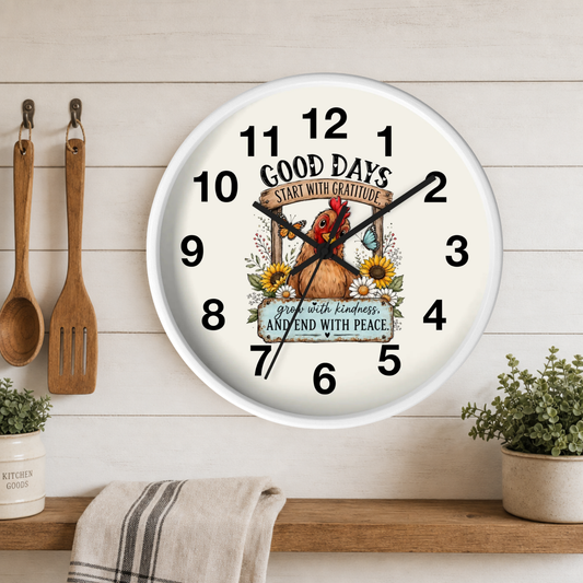 A round wall clock with a white frame and a warm cream face, featuring a decorative illustration of a brown rooster surrounded by sunflowers and the text 'GOOD DAYS START WITH GRATITUDE AND END WITH PEACE.' The clock is mounted on a white wood-paneled wall above a wooden shelf with a towel, hanging wooden utensils, and small potted plants.