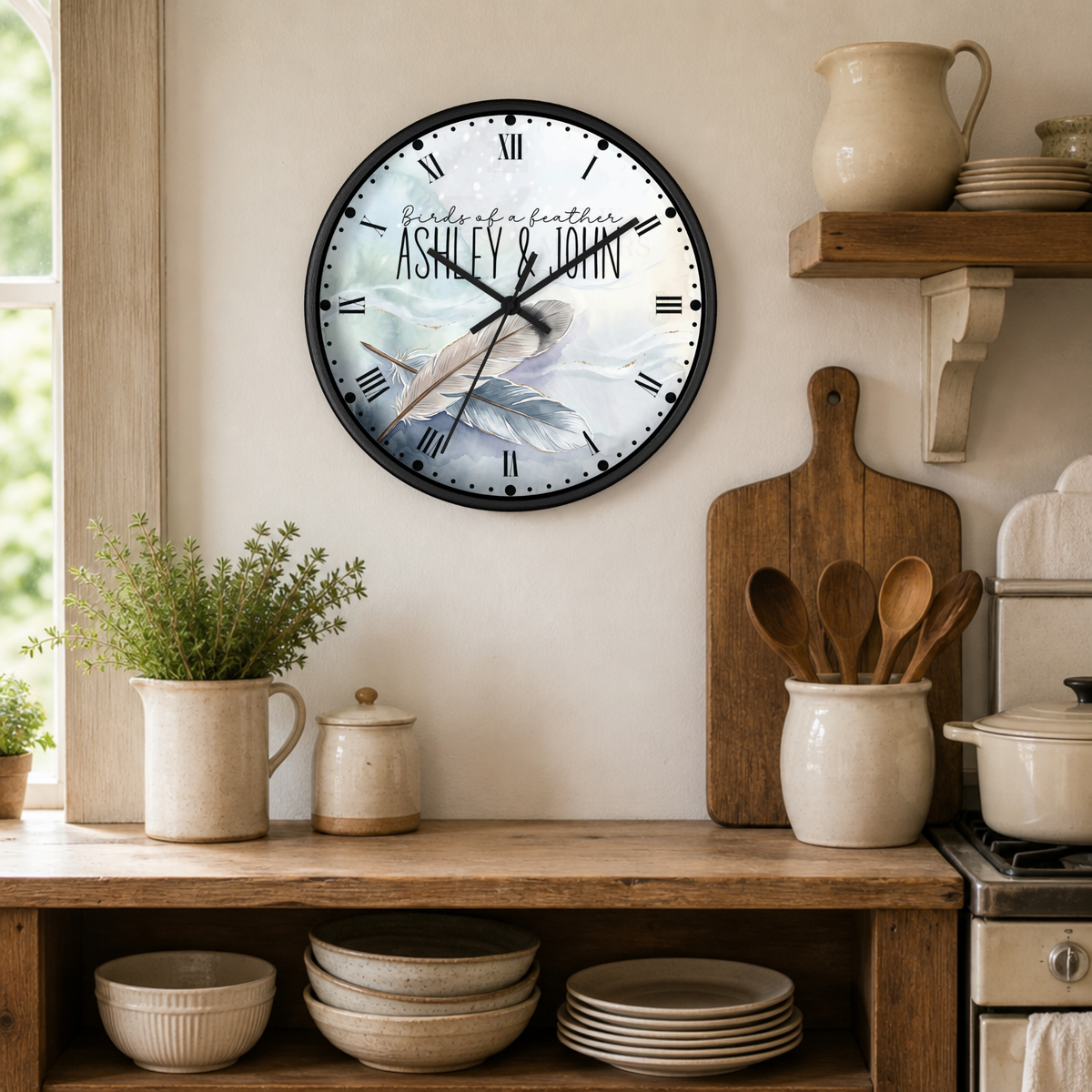 A personalized round wall clock with a black frame hangs on a beige kitchen wall above a wooden counter. The clock face features a bird illustration and the text 'with all love ASHLEY & JOHN' in Roman numerals.