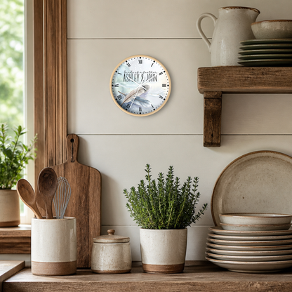 A round wall clock with a rustic wood frame and a printed clock-face design featuring feathers and the Roman numeral XII, mounted on a beige shiplap wall above a wooden shelf with ceramic dishes and plants.