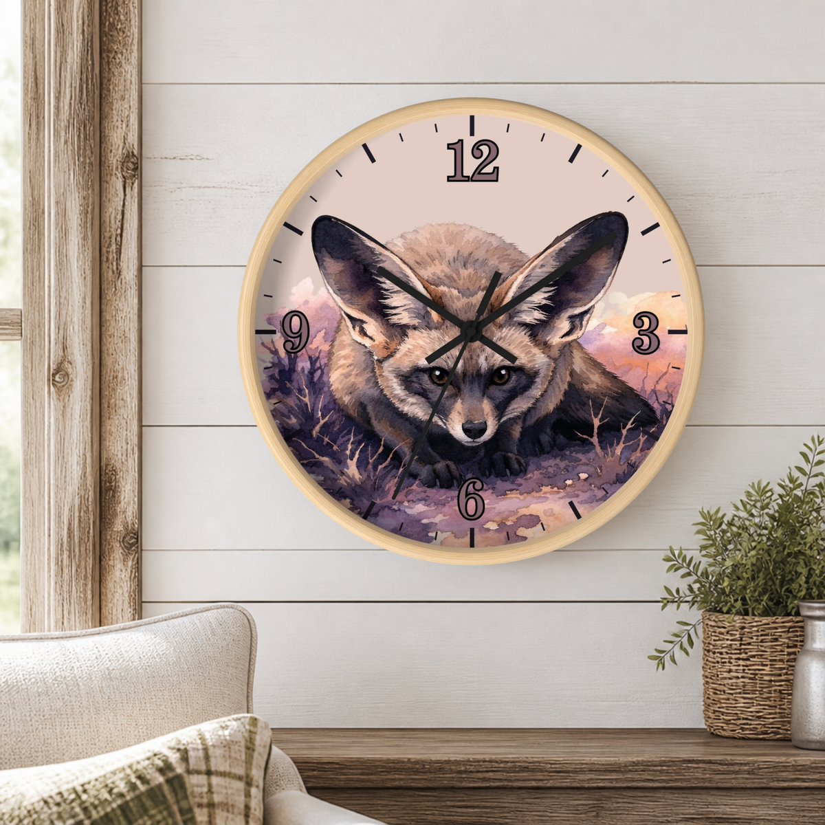A round wall clock with a light wood frame hangs on a white shiplap wall above a wooden mantel. The clock face features a detailed illustration of a large-eared fox in a field at dusk, with black Arabic numerals and black clock hands.