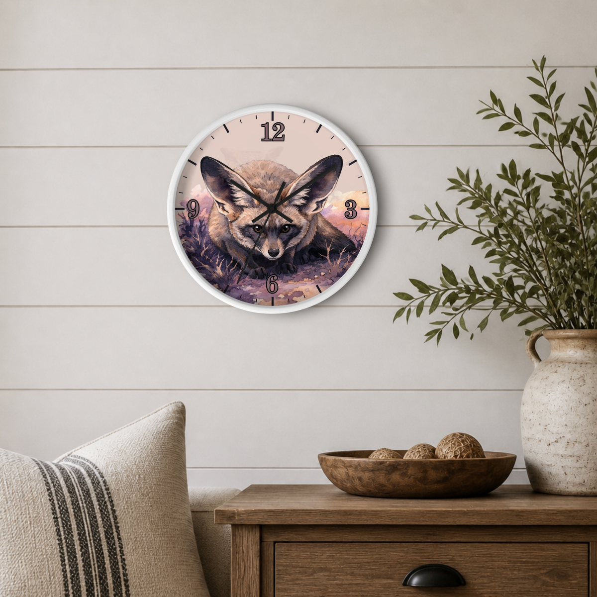 A round wall clock with a white frame hangs on a light wood shiplap wall above a wooden side table. The clock face features a detailed illustration of a fox in natural tones with a sunset sky.