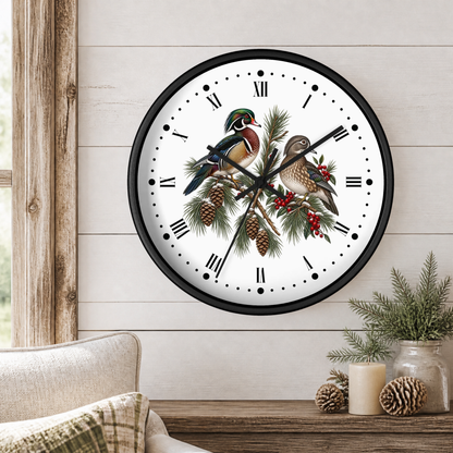 A round wall clock with a black frame and a white face featuring two detailed wood ducks perched on pine branches with pinecones and red berries. The clock is mounted on a light wood-paneled wall above a wooden table with a candle and small plants.
