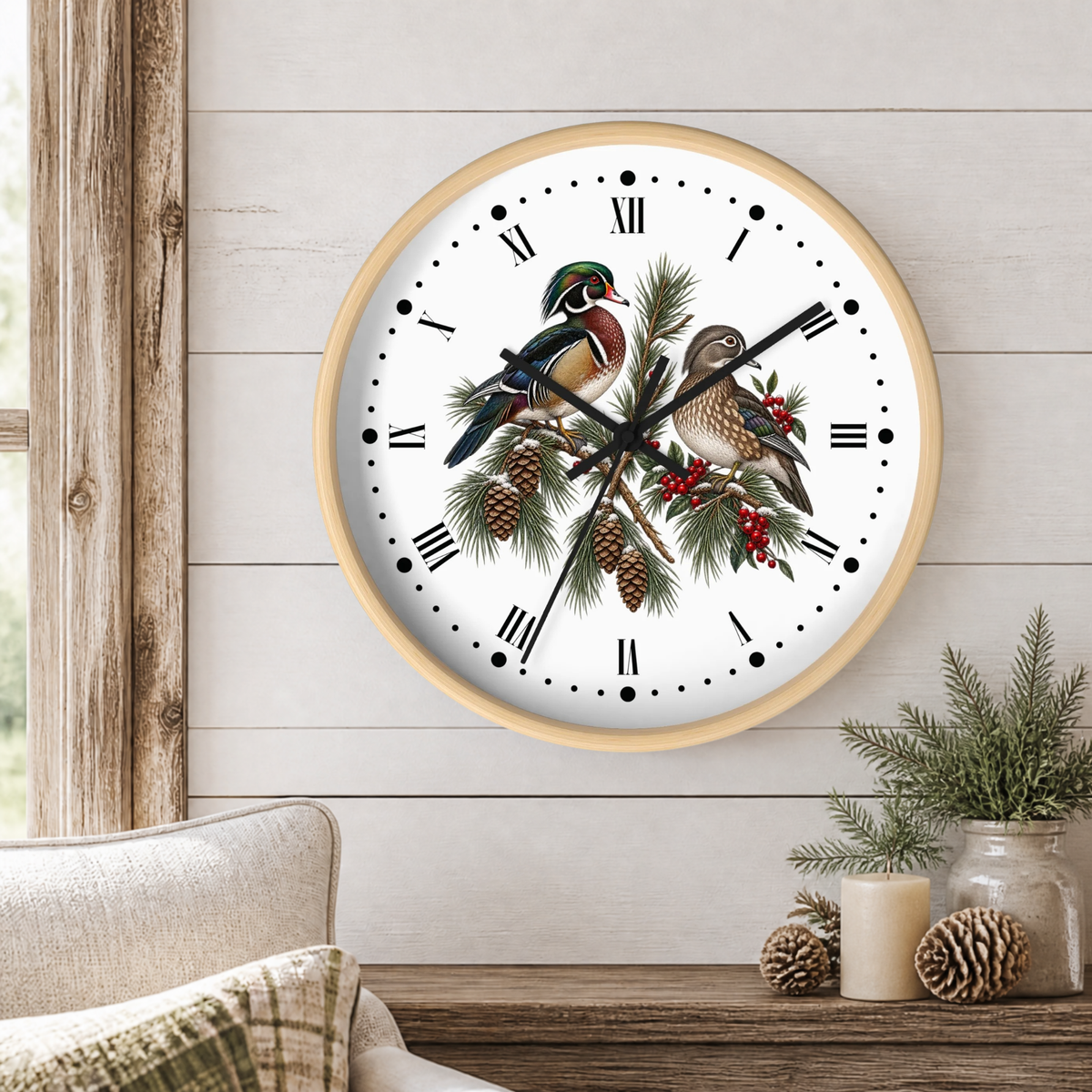 A round wall clock with a light wood frame hangs on a white shiplap wall above a rustic wooden table. The clock face features a white background with black Roman numerals and hands, overlaid with an illustration of two wood ducks perched on pine branches with pinecones and red berries.