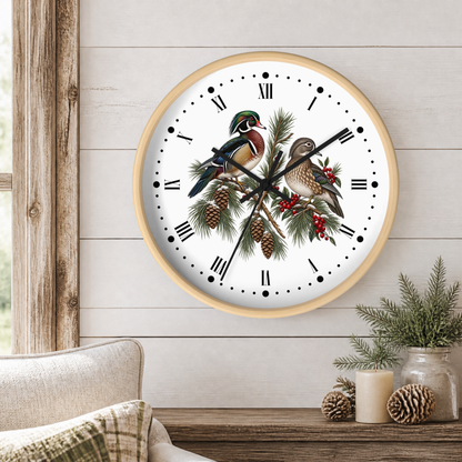 A round wall clock with a light wood frame hangs on a white shiplap wall above a rustic wooden table. The clock face features a white background with black Roman numerals and hands, overlaid with an illustration of two wood ducks perched on pine branches with pinecones and red berries.