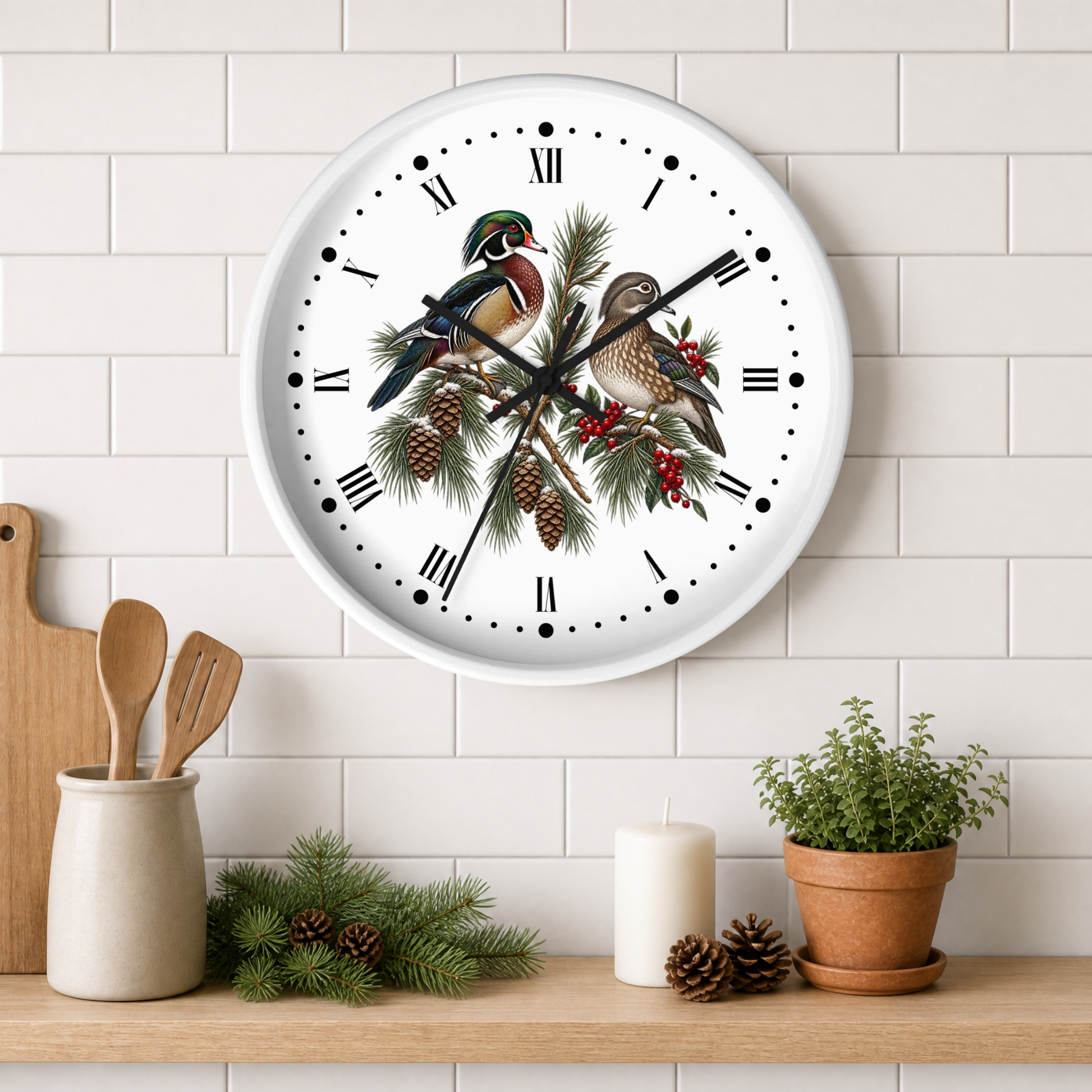 A round white wall clock with a detailed nature illustration of two birds perched on a pine branch with pinecones and red berries. The clock is mounted on a white subway tile wall above a wooden shelf styled with kitchen utensils, a candle, pinecones, and a potted plant.