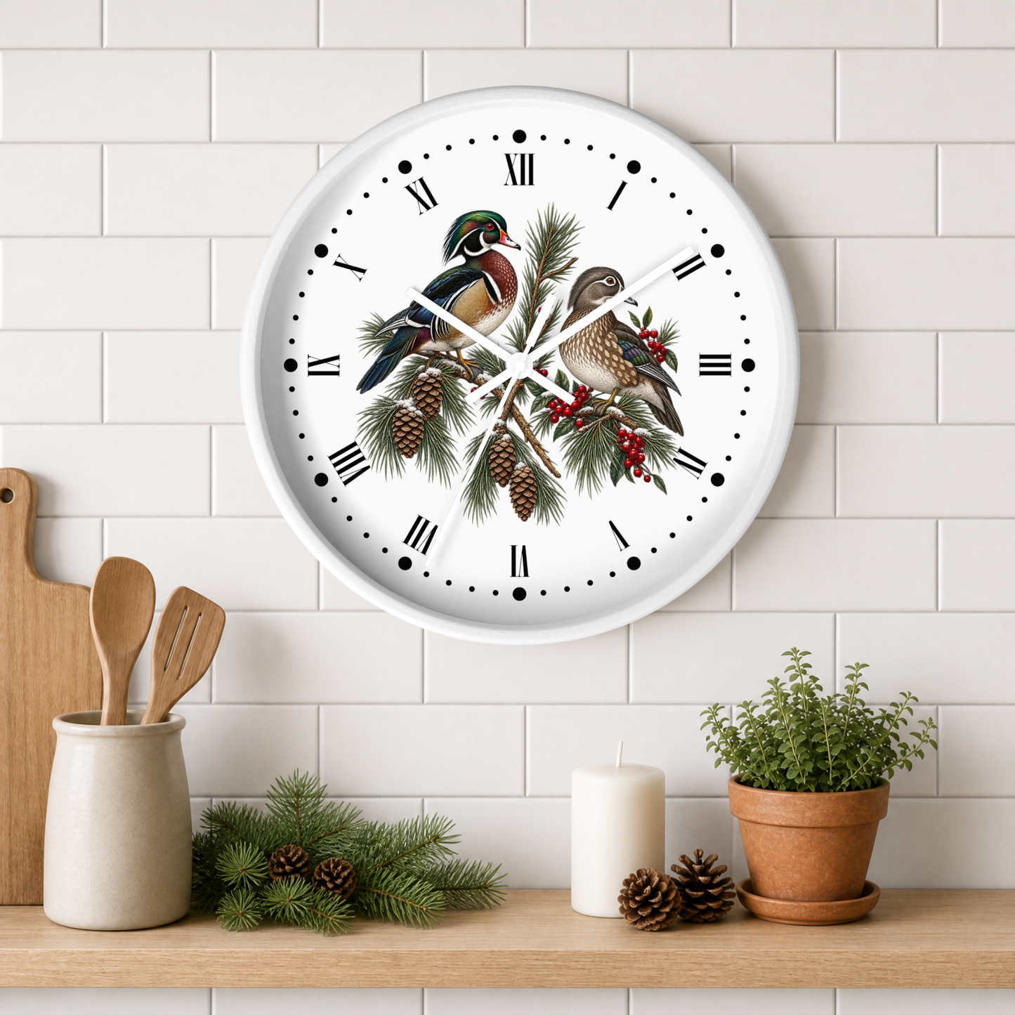 A round wall clock with a white face and Roman numerals, decorated with two wood ducks perched on pine branches with red berries and pinecones. The clock is mounted on a white subway tile wall above a wooden shelf styled with kitchen utensils, candles, pinecones, and a potted plant.