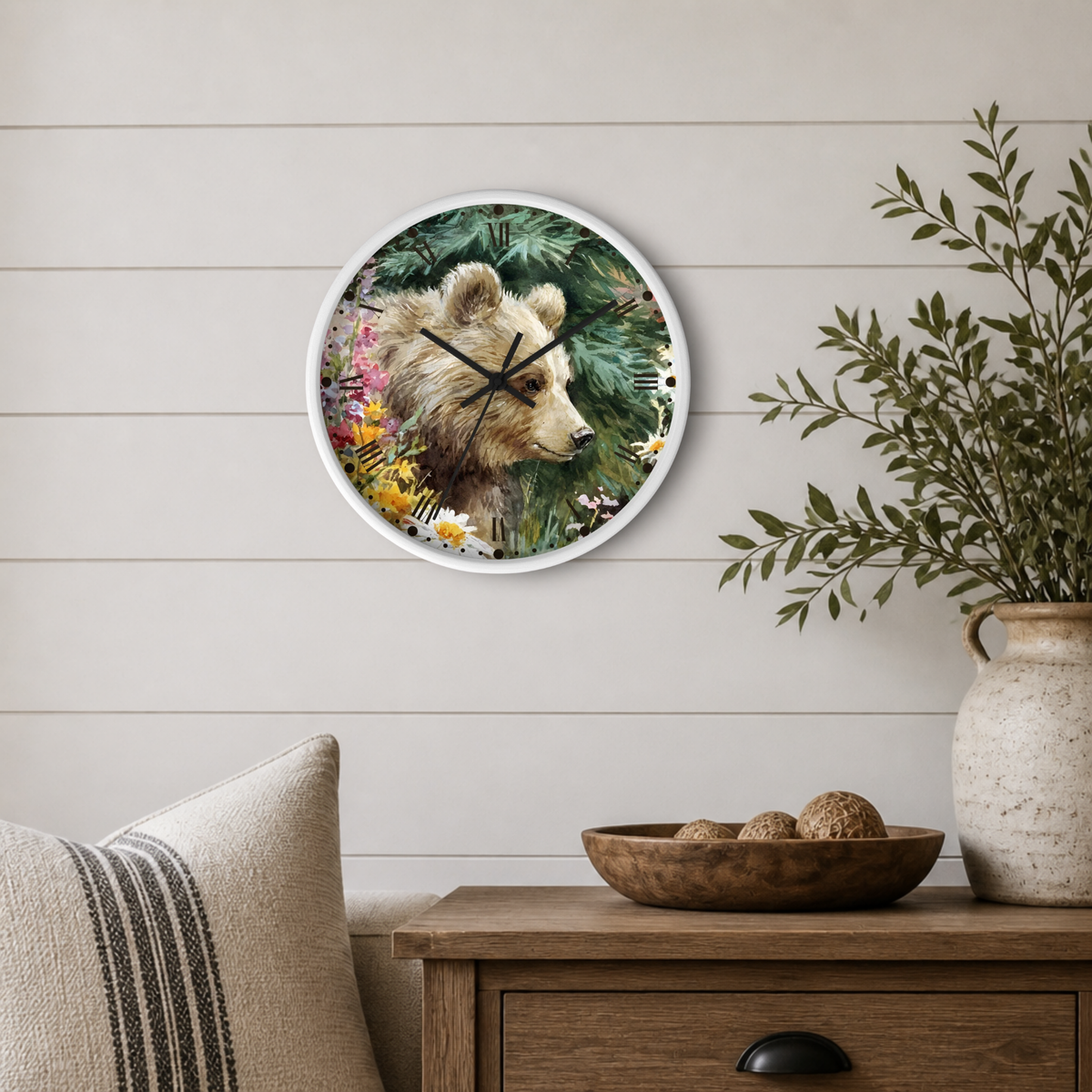 Round wall clock with a white frame featuring a watercolor-style brown bear surrounded by green foliage and colorful wildflowers, mounted on a light beige shiplap wall above a wooden side table with a plant and decorative bowl.