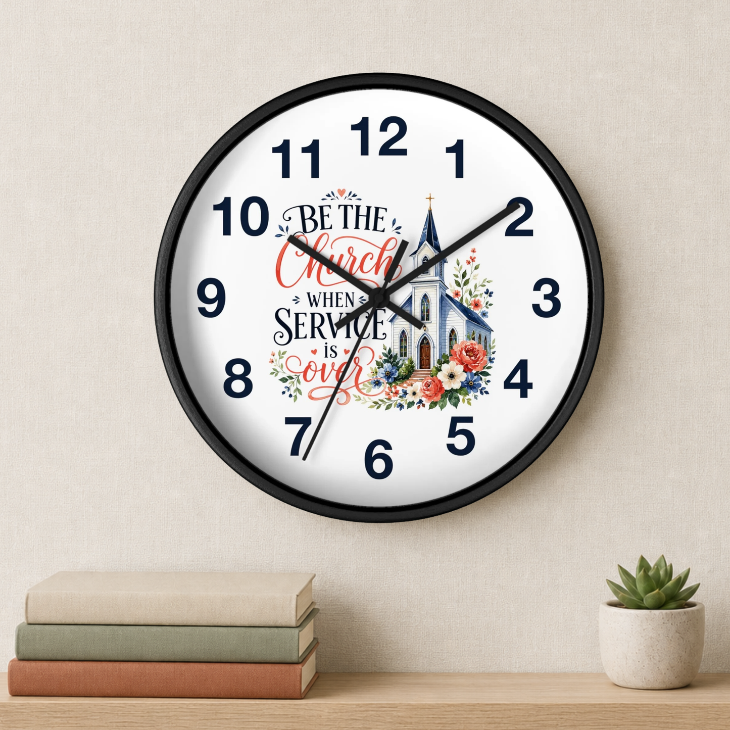 A round wall clock with a black frame and a white face, featuring the text 'Be the Church when Service is over' with a church illustration and floral accents. The clock is mounted on a beige wall above a wooden shelf with stacked books and a small potted succulent.