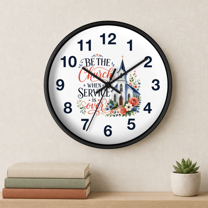 A round wall clock with a black frame and a white face, featuring the text 'Be the Church when Service is over' with a church illustration and floral accents. The clock is mounted on a beige wall above a wooden shelf with stacked books and a small potted succulent.