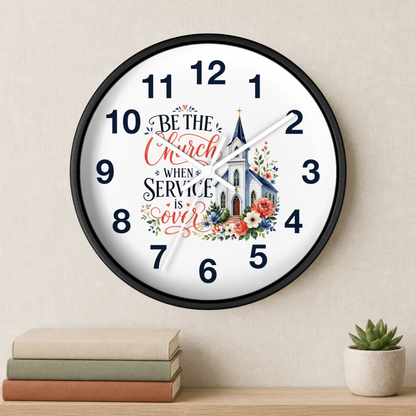 A round wall clock with a black frame and a white face, featuring the text 'BE THE Church WHEN SERVICE IS over' in decorative script, a church illustration, and floral accents. The clock is shown hanging on a beige wall above a shelf with stacked books and a small potted succulent.
