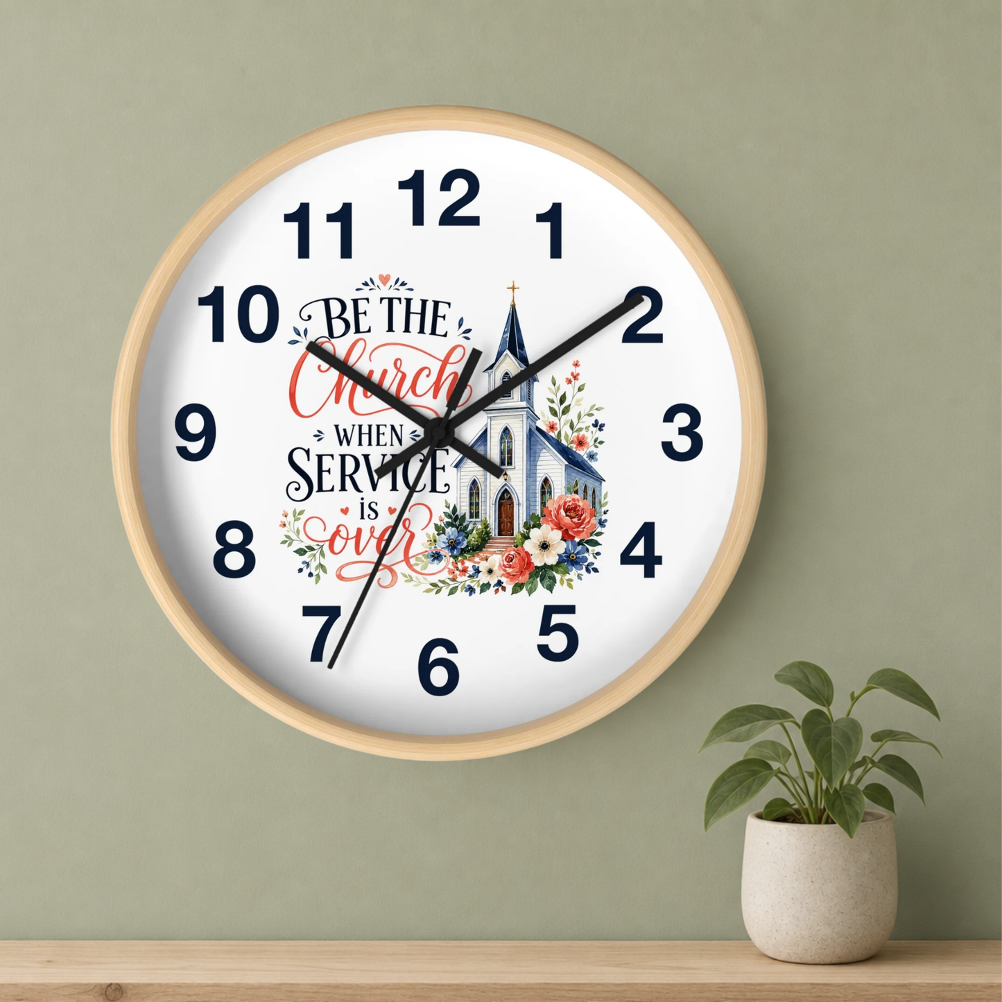 A round wall clock with a light wood frame hangs on a sage green wall above a wooden shelf. A small potted green plant sits on the shelf to the right.