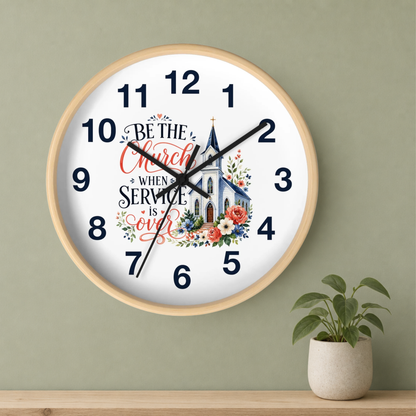 A round wall clock with a light wood frame hangs on a sage green wall above a wooden shelf. A small potted green plant sits on the shelf to the right.