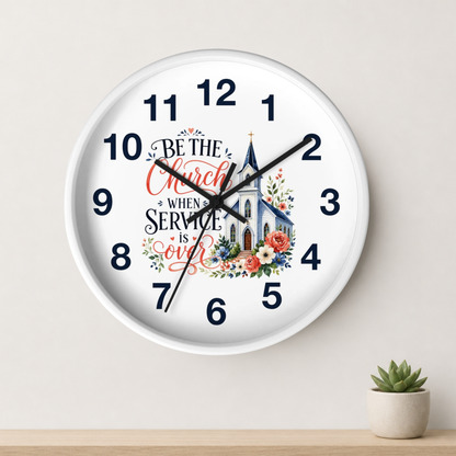 A round white wall clock with black numerals and hands, featuring a church illustration and the phrase 'Be the Church When Service is Over' in red and black script. The clock is mounted on a light beige wall above a wooden shelf with a small potted succulent.