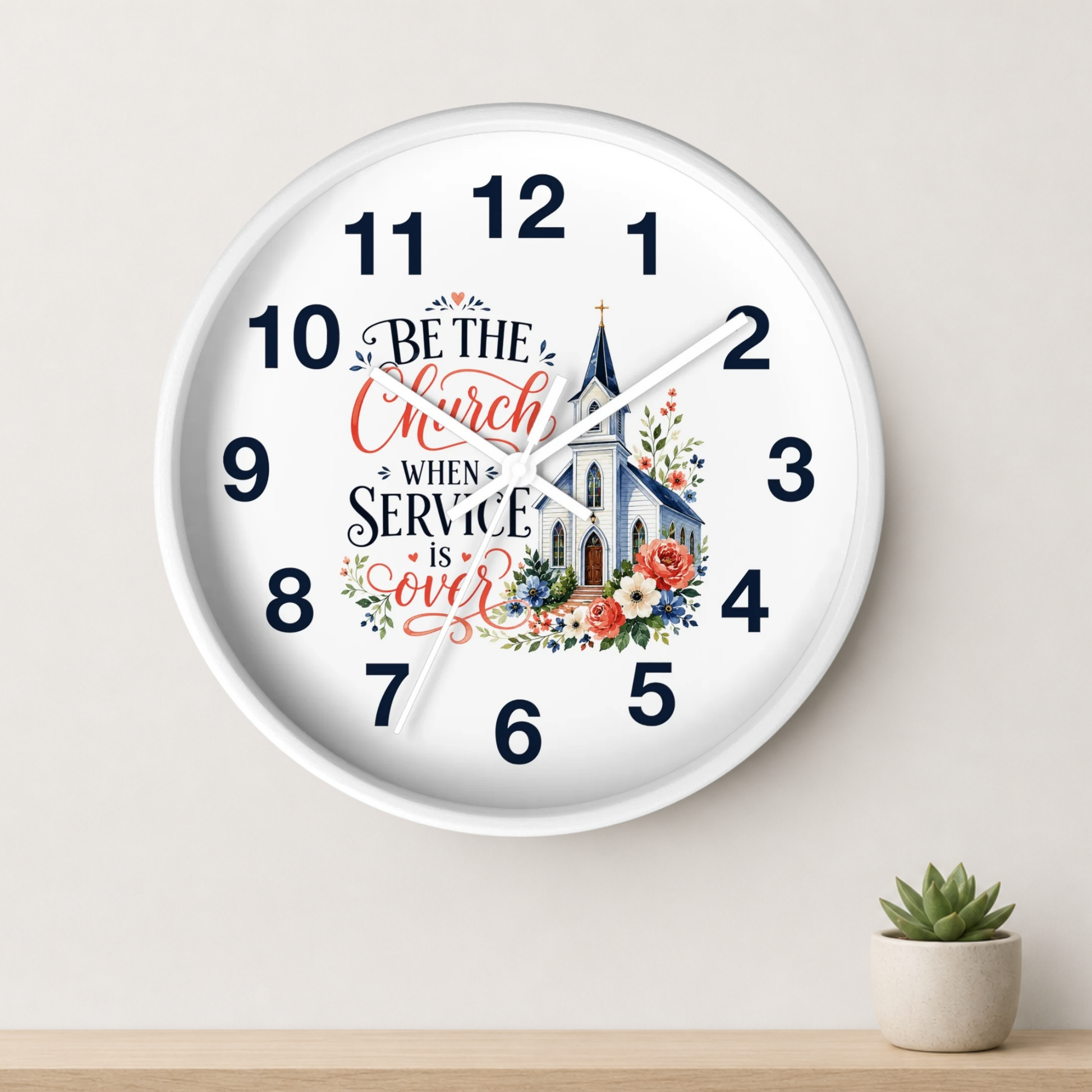 A round white wall clock with navy numerals and hands, featuring a church illustration and the phrase 'Be the Church When Service Is Over' in decorative script. The clock is mounted on a light beige wall above a wooden surface with a small potted succulent.