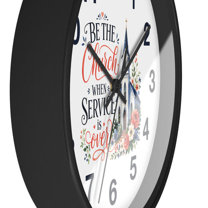 A close-up of a round wall clock with a black frame and a white face featuring the phrase 'Be the Church When Service is Over' in black and red script, alongside a church illustration and floral details.