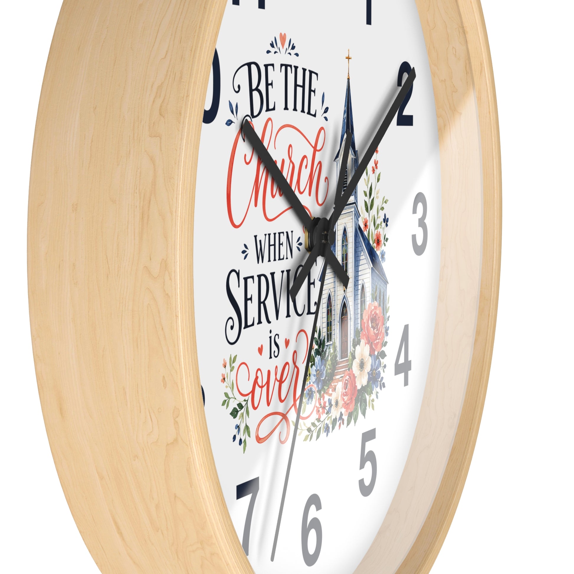 A close-up of a round wall clock with a light natural wood frame and a white face. The clock features black hands, black numerals, and a decorative script quote reading 'Be the Church when service is over' in red and black lettering, accompanied by a church illustration and floral accents.