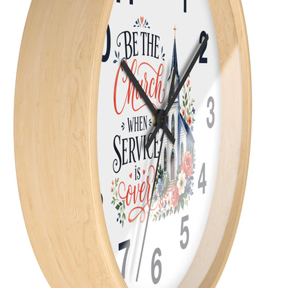 A close-up of a round wall clock with a light natural wood frame and a white face. The clock features black hands, black numerals, and a decorative script quote reading 'Be the Church when service is over' in red and black lettering, accompanied by a church illustration and floral accents.