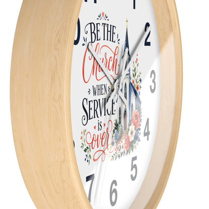 A close-up of a round wall clock with a light natural wood frame and a white face. The clock face features a church illustration and the hand-lettered quote, 'BE THE Church WHEN SERVICE is over' in black and coral-red script, with floral accents.