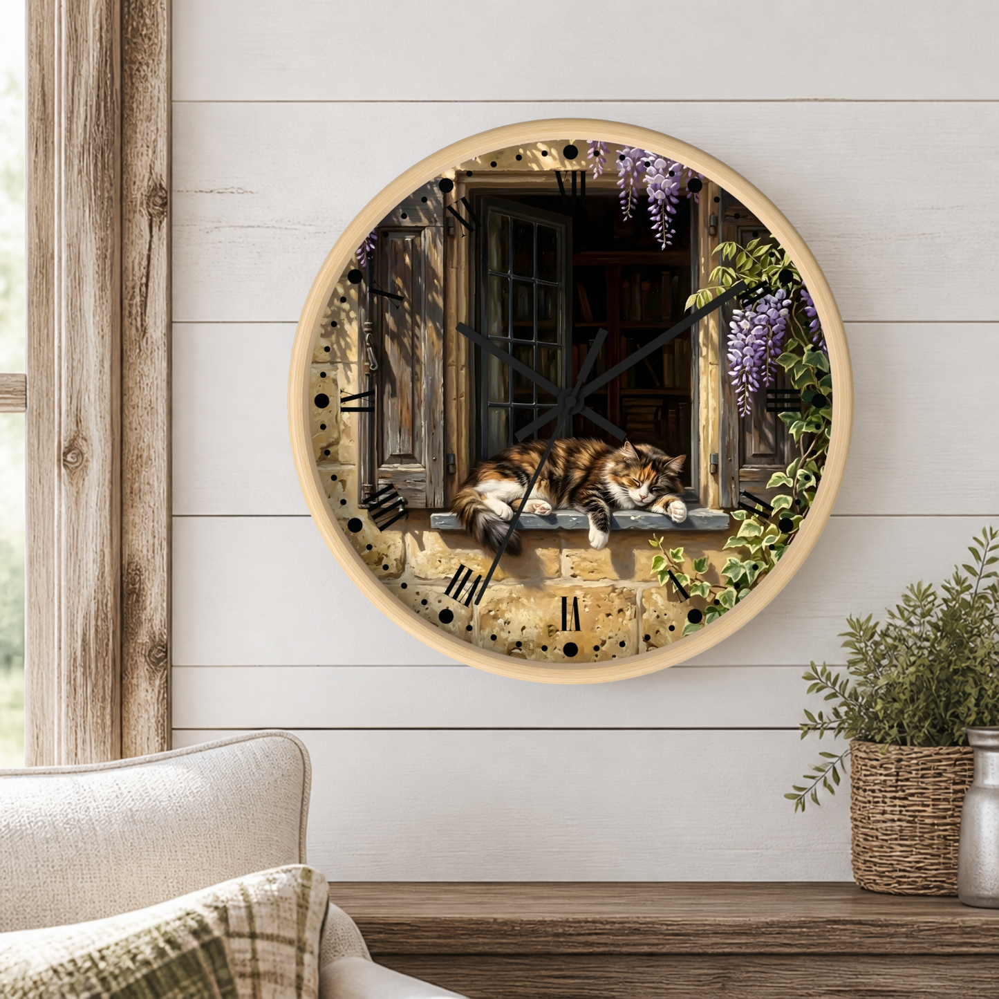 Round wall clock with a light wood frame, featuring a printed design of a brown calico cat sleeping on a window sill with purple wisteria flowers, mounted on a white shiplap wall above a wooden mantel with a plant.