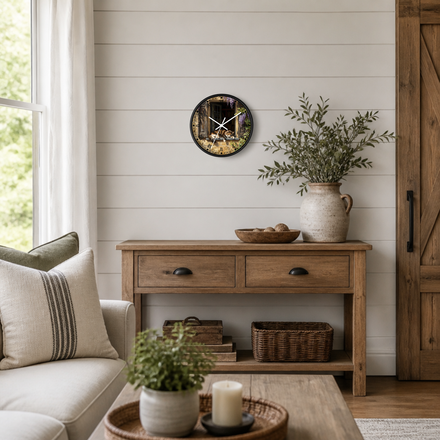 A round wall clock with a lounging calico cat scene print hangs on a white shiplap wall above a wooden console table in a styled living room.