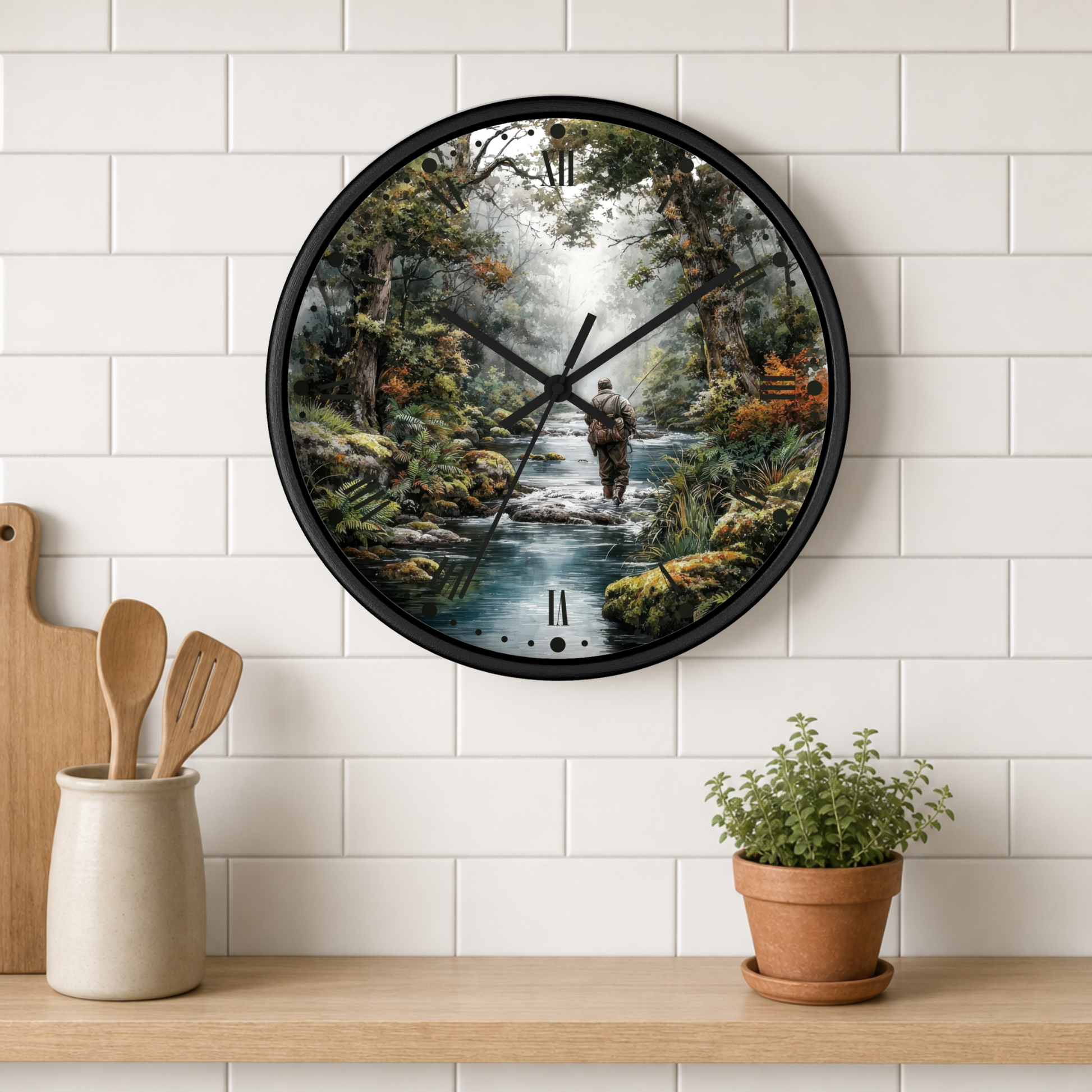 A round wall clock with a black frame and a printed forest scene featuring a fly-fishing figure, mounted on a white subway tile wall above a wooden shelf with kitchen utensils and a potted plant.