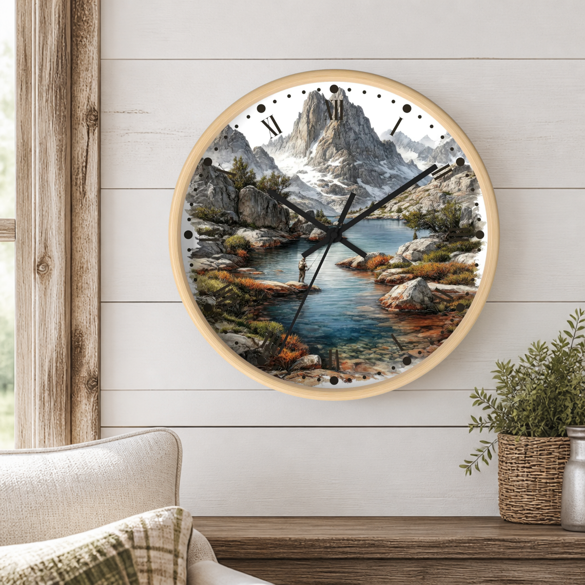 Round wall clock with a light wood frame and a scenic mountain river landscape print, mounted on a white wood-paneled wall above a wooden table with a plant and part of a chair visible.