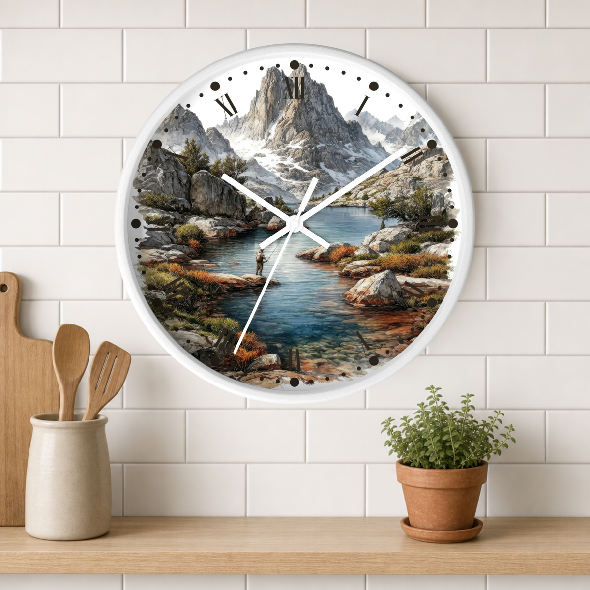 A round wall clock with a scenic mountain lake print, hanging on a white subway tile wall above a wooden shelf with a small potted plant and wooden utensils.