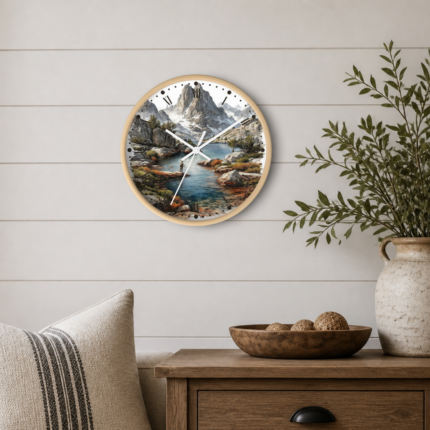 Round wall clock with a natural wood frame and a scenic mountain lake print, mounted on a white shiplap wall above a wooden side table with a plant and decorative bowl.