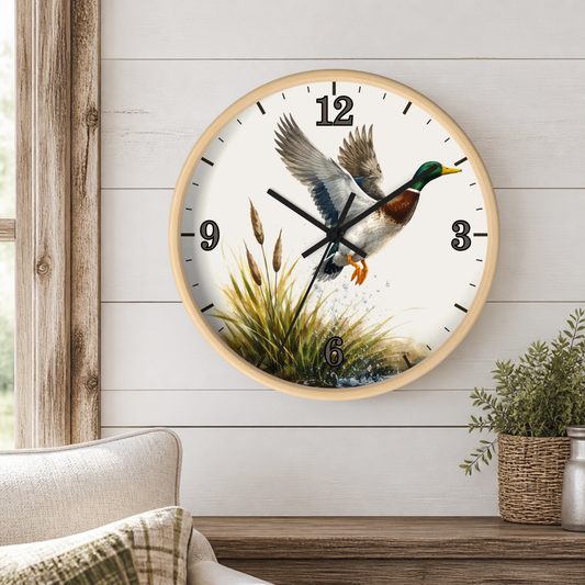 A round wall clock with a light wood frame hangs on a white shiplap wall above a wooden mantel. The clock face features a detailed illustration of a mallard duck in flight over reeds, with black Arabic numerals and black clock hands.