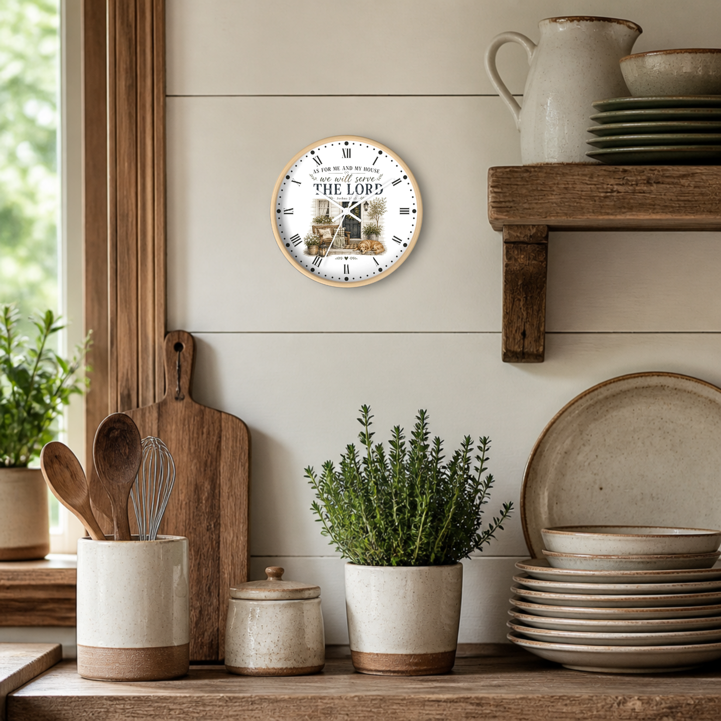 A round wall clock with a light wood frame hangs on a beige shiplap wall in a rustic kitchen setting. The clock face features a scenic illustration of a cottage with the text 'For Me And My House We Will Serve THE LORD' 