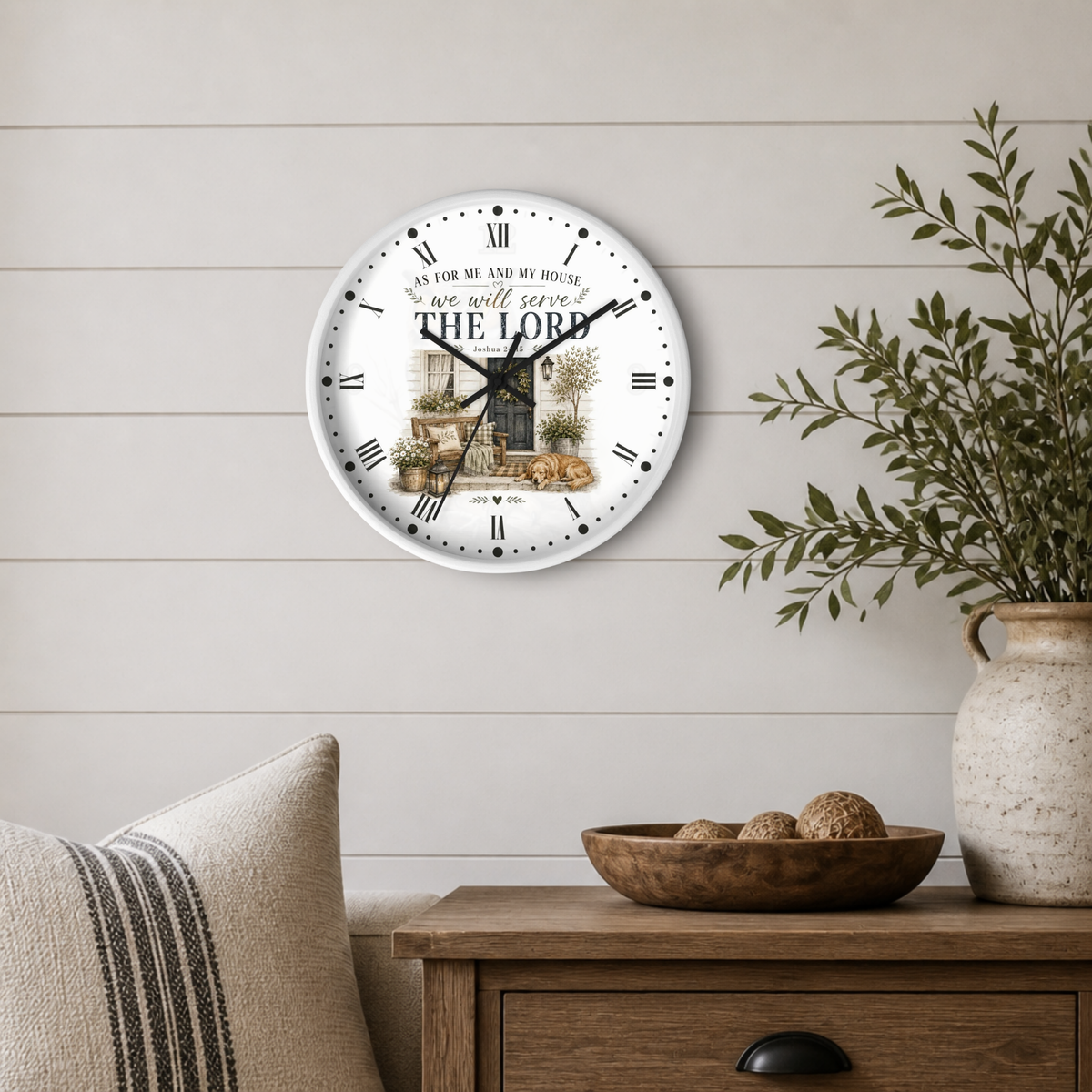 A round wall clock with a white face, black Roman numerals, and a rustic illustration of a garden scene with the text 'We will always THE LORD'. The clock is mounted on a light beige shiplap wall above a wooden side table with a vase of greenery and a striped pillow.