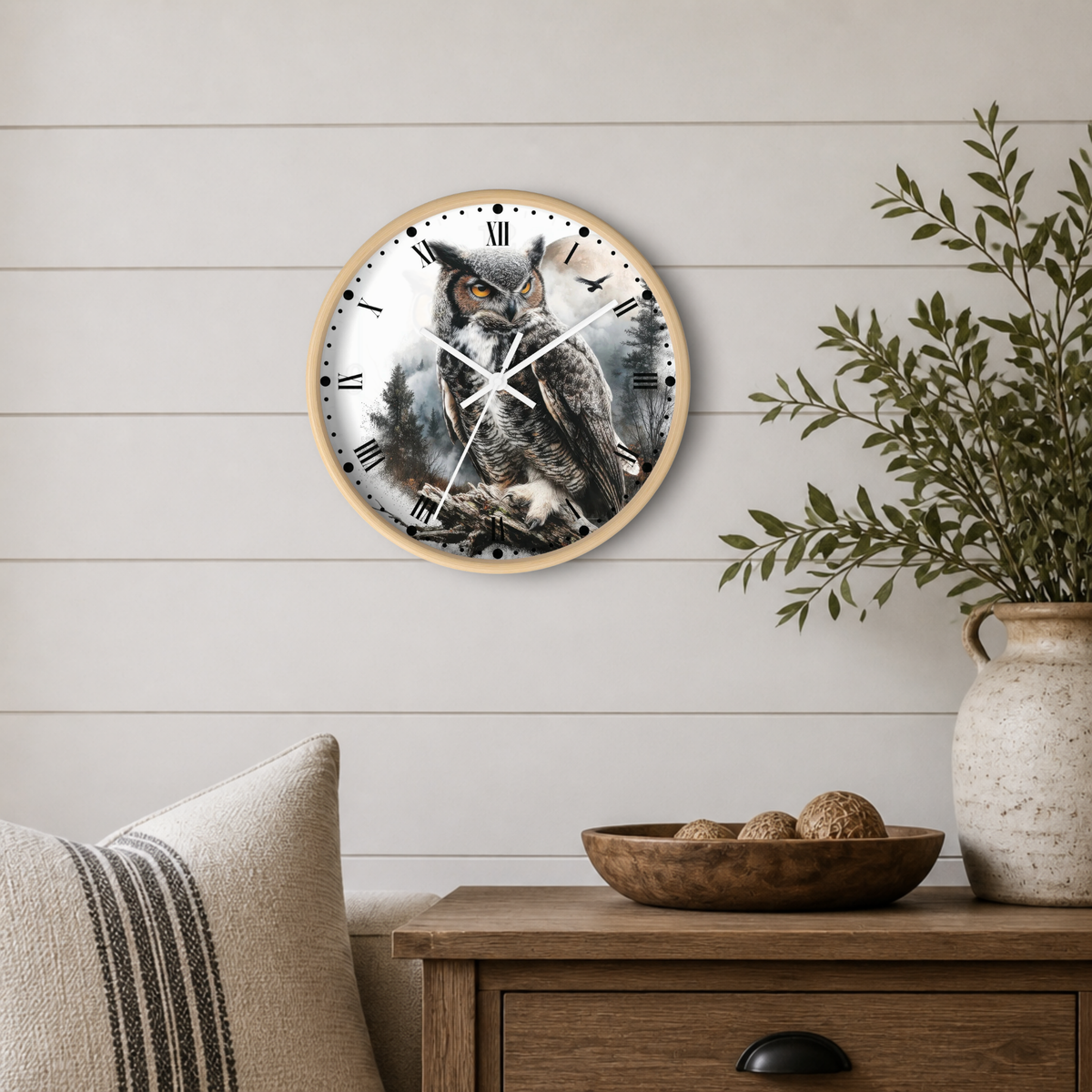 A round wall clock with a natural wood frame and a detailed owl illustration on the face, mounted on a white shiplap wall above a wooden side table with a plant and decorative bowl.