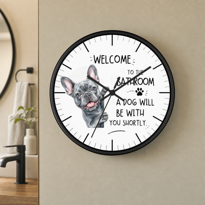 A round wall clock with a black frame and a white face featuring a French bulldog illustration and the text 'WELCOME TO THE BATHROOM A DOG WILL BE WITH YOU SHORTLY.' The clock is mounted on a beige wall in a bathroom setting.