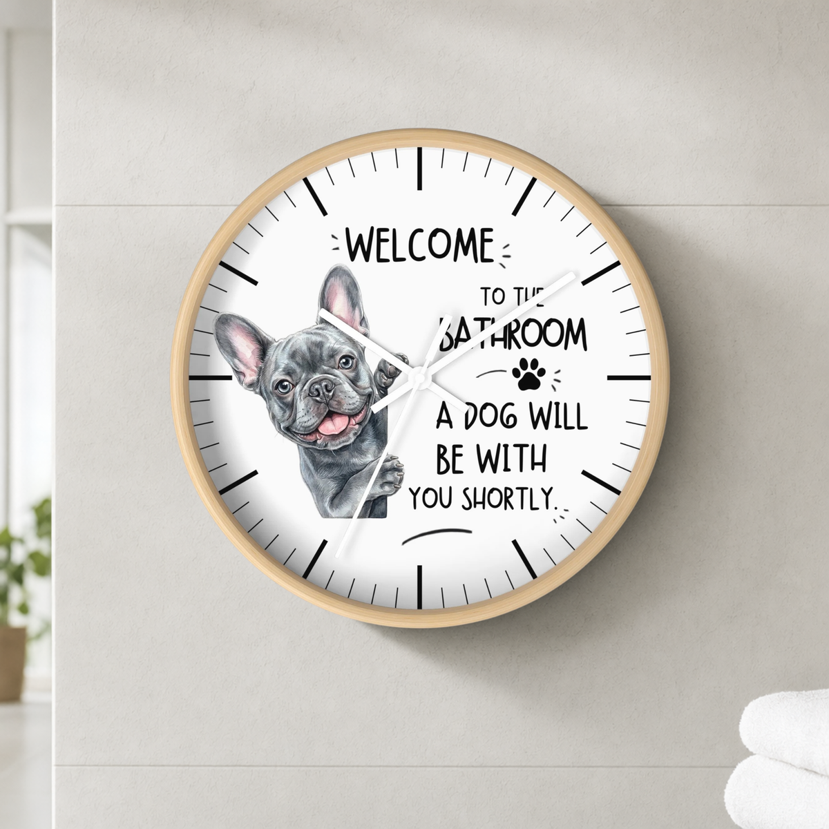 A round wall clock with a light wood frame and a white face, mounted on a light gray tiled wall. The clock features a black and white illustration of a French Bulldog and the humorous text 'WELCOME TO THE BATHROOM A DOG WILL BE WITH YOU SHORTLY.