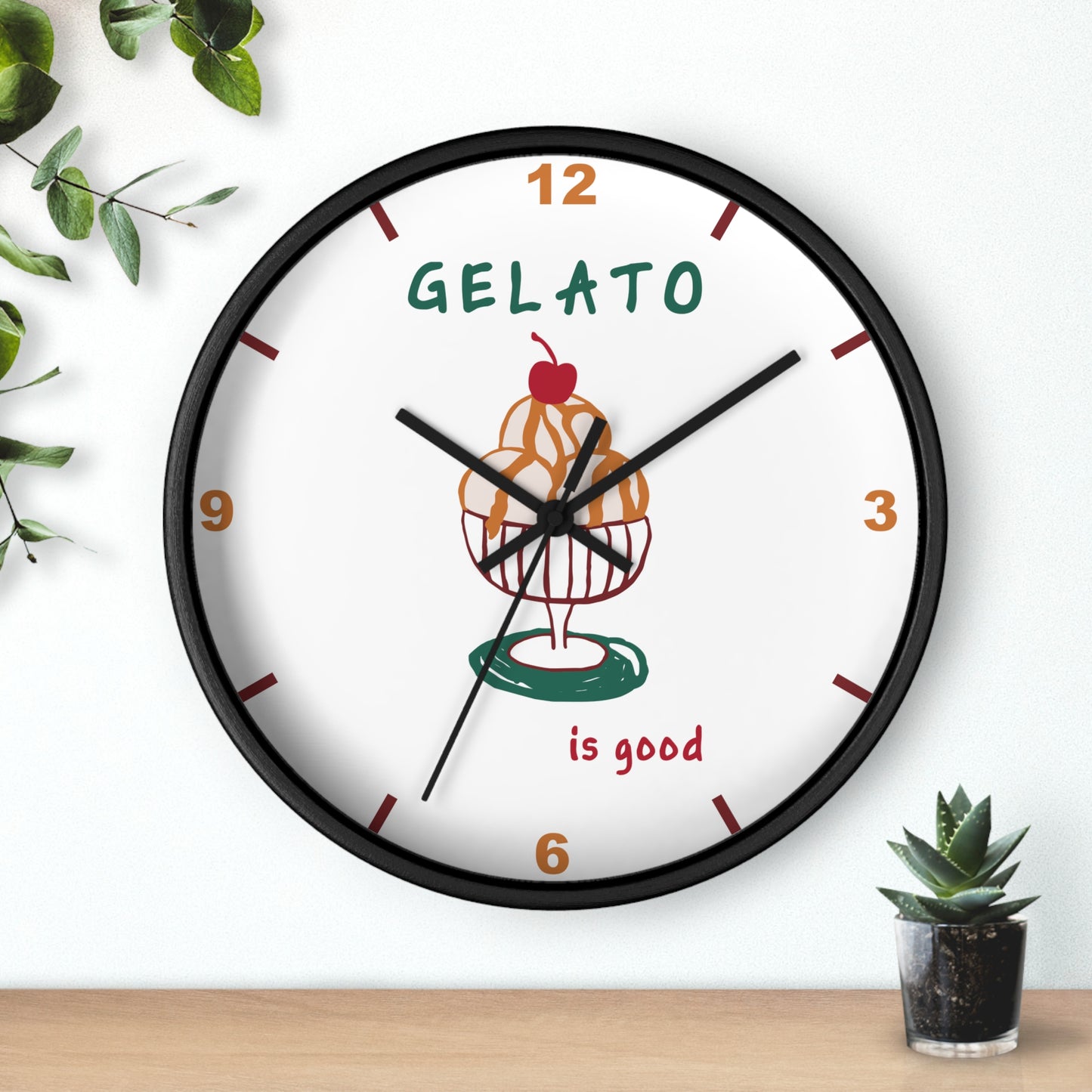 A round wall clock with a black frame and a white face, featuring a gelato cupcake illustration and the words 'GELATO is good'. The clock is mounted on a white wall above a light wood surface, with decorative green leaves and a small succulent nearby.
