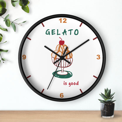 A round wall clock with a black frame and a white face, featuring a gelato cupcake illustration and the words 'GELATO is good'. The clock is mounted on a white wall above a light wood surface, with decorative green leaves and a small succulent nearby.