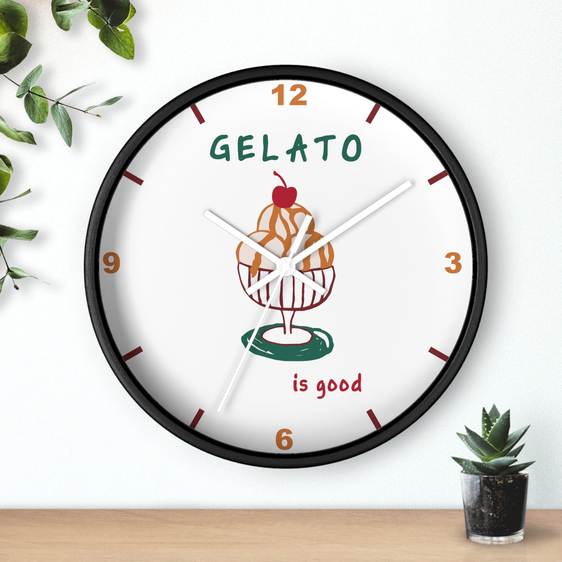A round wall clock with a black frame and a white face, featuring a gelato cupcake illustration and the words 'GELATO is good'. The clock is mounted on a white wall above a light wood surface, with decorative green leaves and a small succulent nearby.